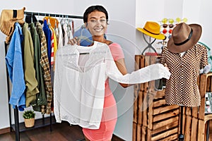 Young hispanic woman customer smiling confident choosing clothes holding shirt at clothing store