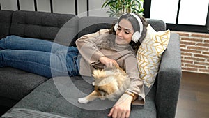 Young hispanic woman with dog listening to music lying on sofa at home