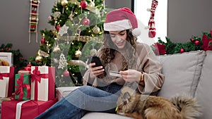 Young hispanic woman with dog shopping with smartphone and credit card celebrating christmas at home