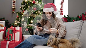 Young hispanic woman with dog shopping with smartphone and credit card celebrating christmas at home