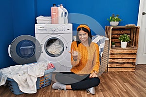 Young hispanic woman doing laundry beckoning come here gesture with hand inviting welcoming happy and smiling