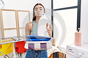 Young hispanic woman doing laundry holding detergent bottle and folded clothes puffing cheeks with funny face