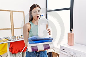 Young hispanic woman doing laundry holding detergent bottle and folded clothes in shock face, looking skeptical and sarcastic,