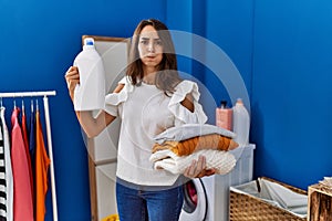 Young hispanic woman doing laundry holding detergent bottle puffing cheeks with funny face