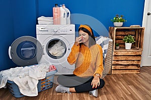 Young hispanic woman doing laundry smelling something stinky and disgusting, intolerable smell, holding breath with fingers on