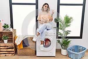 Young hispanic woman drinking coffee waiting for washing machine at laundry room smiling with happy face winking at the camera