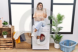 Young hispanic woman drinking coffee waiting for washing machine at laundry room very happy and excited doing winner gesture with