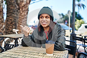 Young hispanic woman drinking a cup of coffee outdoors pointing thumb up to the side smiling happy with open mouth