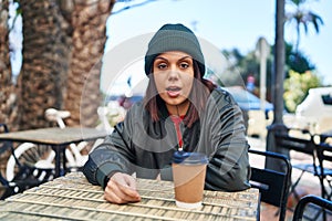 Young hispanic woman drinking a cup of coffee outdoors scared and amazed with open mouth for surprise, disbelief face