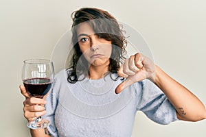 Young hispanic woman drinking a glass of red wine with angry face, negative sign showing dislike with thumbs down, rejection