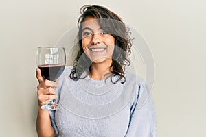 Young hispanic woman drinking a glass of red wine looking positive and happy standing and smiling with a confident smile showing