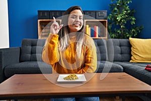 Young hispanic woman eating green peas smiling with an idea or question pointing finger up with happy face, number one