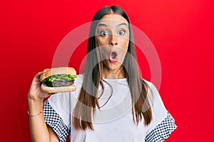 Young hispanic woman eating hamburger scared and amazed with open mouth for surprise, disbelief face