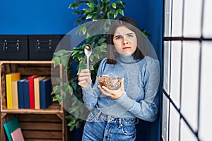 Young hispanic woman eating healthy whole grain cereals with spoon clueless and confused expression