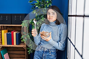 Young hispanic woman eating healthy whole grain cereals with spoon puffing cheeks with funny face
