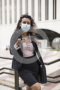 Young hispanic woman with face mask using her smart phone outdoors. Enterprising person