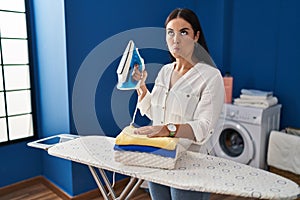 Young hispanic woman with folded laundry after ironing making fish face with mouth and squinting eyes, crazy and comical