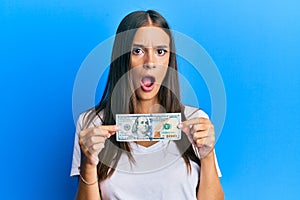 Young hispanic woman holding 100 dollar banknote in shock face, looking skeptical and sarcastic, surprised with open mouth