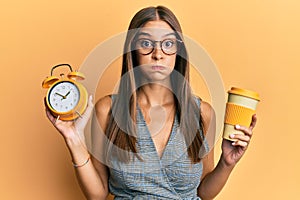 Young hispanic woman holding alarm clock and drinking a take away cup of coffee puffing cheeks with funny face