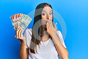 Young hispanic woman holding australian dollars covering mouth with hand, shocked and afraid for mistake