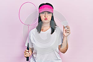 Young hispanic woman holding badminton racket and shuttlecock puffing cheeks with funny face
