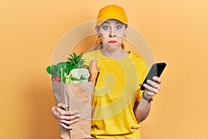 Young hispanic woman holding bag of groceries using smartphone puffing cheeks with funny face