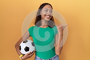 Young hispanic woman holding ball smiling with happy face looking and pointing to the side with thumb up