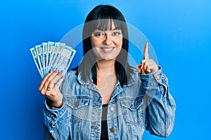 Young hispanic woman holding bangladesh taka banknotes smiling with an idea or question pointing finger with happy face, number