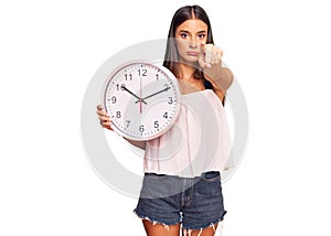 Young hispanic woman holding big clock pointing with finger to the camera and to you, confident gesture looking serious