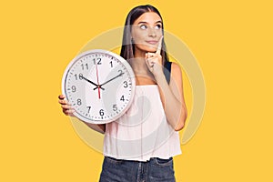Young hispanic woman holding big clock serious face thinking about question with hand on chin, thoughtful about confusing idea