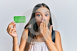 Young hispanic woman holding birth control pills covering mouth with hand, shocked and afraid for mistake