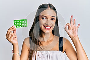 Young hispanic woman holding birth control pills doing ok sign with fingers, smiling friendly gesturing excellent symbol