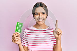 Young hispanic woman holding birth control pills smiling with an idea or question pointing finger with happy face, number one