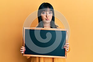 Young hispanic woman holding blackboard puffing cheeks with funny face