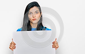 Young hispanic woman holding blank empty banner thinking attitude and sober expression looking self confident