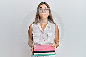 Young hispanic woman holding books puffing cheeks with funny face