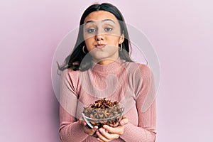 Young hispanic woman holding bowl of star anise puffing cheeks with funny face