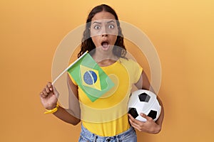 Young hispanic woman holding brazil flag and football ball in shock face, looking skeptical and sarcastic, surprised with open
