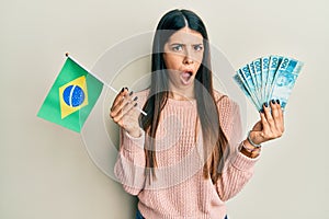 Young hispanic woman holding brazil flag and real banknotes in shock face, looking skeptical and sarcastic, surprised with open