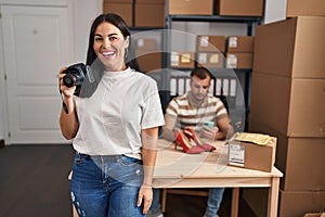 Young hispanic woman holding camera working at small business ecommerce looking positive and happy standing and smiling with a