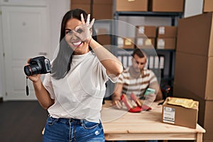 Young hispanic woman holding camera working at small business ecommerce smiling happy doing ok sign with hand on eye looking