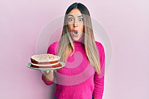 Young hispanic woman holding carrot cake scared and amazed with open mouth for surprise, disbelief face