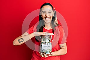 Young hispanic woman holding charity jar with money smiling and laughing hard out loud because funny crazy joke