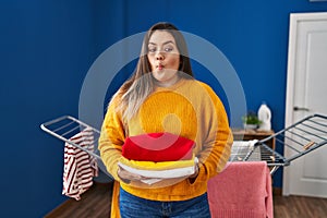 Young hispanic woman holding clean and folded laundry making fish face with mouth and squinting eyes, crazy and comical