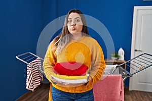 Young hispanic woman holding clean and folded laundry puffing cheeks with funny face