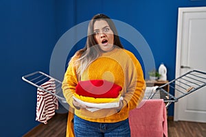 Young hispanic woman holding clean and folded laundry in shock face, looking skeptical and sarcastic, surprised with open mouth