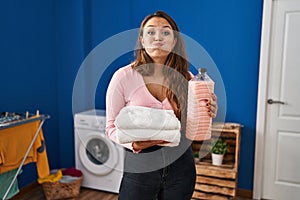 Young hispanic woman holding clean laundry and detergent bottle puffing cheeks with funny face