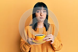 Young hispanic woman holding coffee puffing cheeks with funny face