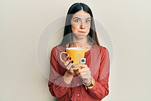 Young hispanic woman holding cup of tea puffing cheeks with funny face