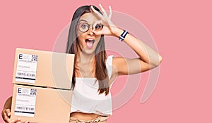 Young hispanic woman holding delivery package smiling happy doing ok sign with hand on eye looking through fingers